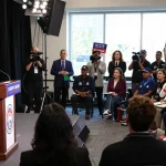 Casey Askar debating in a packed audience during a political campaign highlighting his controversies.