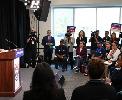 Casey Askar debating in a packed audience during a political campaign highlighting his controversies.